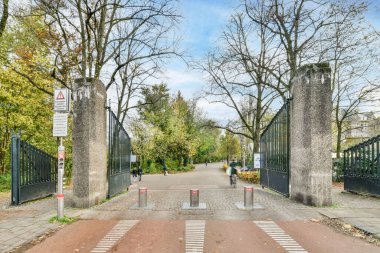 Amsterdam, Hollanda - 10 Nisan 2021: Arka planda ağaçlar ve beyaz bulutlarla kaplı mavi gökyüzü olan bir parka giriş