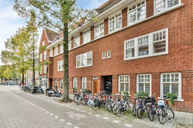 Amsterdam, Hollanda - 10 Nisan 2021: Beyaz süslemeli ve pencereli kırmızı tuğlalı bir binanın önündeki caddenin kenarına parketmiş bisikletler