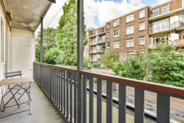 Amsterdam, Hollanda - 10 Nisan 2021: Arka planda sandalyeler ve ağaçlar olan bir balkon, apartman binalarının balkon penceresinden sokağa bakıyor.