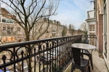 Masası ve sol tarafında sandalyeleri olan bir balkon. Apartman penceresinden sokağa bakıyor.