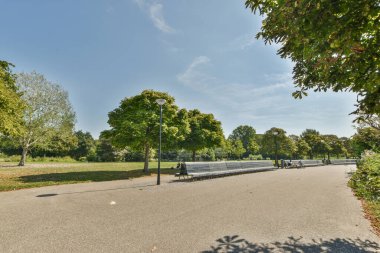 Parktaki gölge ağaçlarının altındaki banklarda yürüyen ve oturan insanların olduğu bir park yeşil yapraklarla doludur.