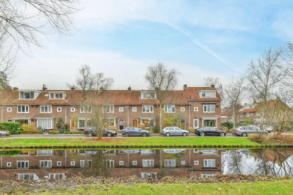 stock image Amsterdam, Netherlands - 10 April, 2021: some houses that are reflected in the water with cars parked on the other side of the house and trees behind them