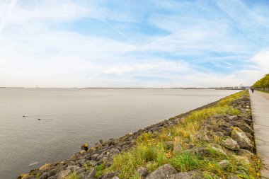 Amsterdam, Hollanda - 10 Nisan 2021: Bir yol kenarındaki su ve kayalar uzaktaki bir tekne ile bir su kütlesine götürür.