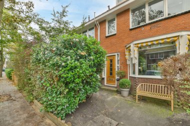 Evin ön bahçesinde bir bank ve evin kenarında büyüyen çalılar olan bir ev. Yukarıda parlak mavi bir gök var.