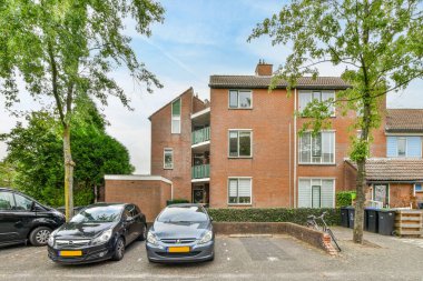 Her iki tarafında ağaçlar ve binaların üstünde mavi bir gökyüzü olan tuğla bir binanın önüne park edilmiş iki araba.