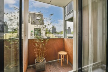 Bir balkon, bir sandalye ve yerde saksı bitkisi, açık bir pencerenin önünde dışarıya bakıyor.