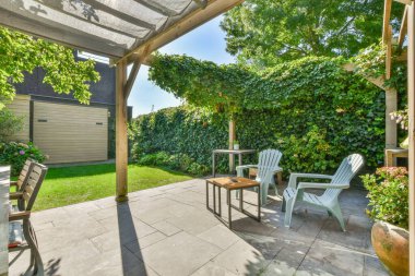 Bahçesinde sandalyeler ve ağaçlar olan, güneşli mavi gökyüzünde yemyeşil yapraklarla çevrili bir bahçe.