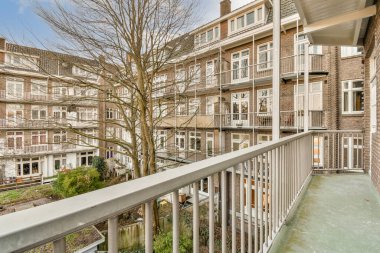 Arka planda ağaçlar ve binalar olan bir dış alan. Balkondan alınmış ve yaprakları olmayan bir ağaca bakıyor.