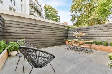 Arka bahçesinde ağaçlar, ağaçlar ve çalılar olan sandalyeler ve masalar.