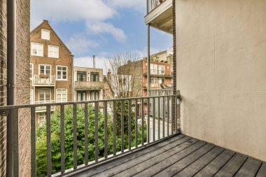 Arka planda ağaçlar ve binalar olan bir balkon. Bir apartmanın penceresinden sokağa bakıyor.