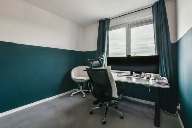 Masası, sandalyesi ve bilgisayar monitörü olan bir oda şehre bakan bir pencerenin önünde.