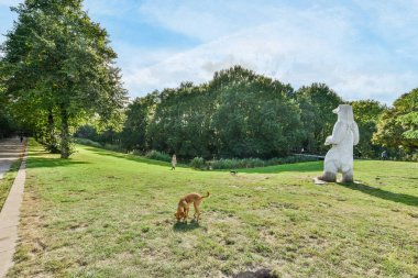 Amsterdam, Hollanda - 10 Nisan 2021: Güneşli bir günde büyük bir taş heykelin yanında ağaçlar ve çalılar olan bir köpek duruyor.