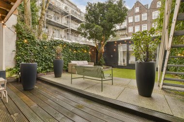 Binanın yan tarafında ağaçlar, çimenler ve çalılarla çevrili sandalye ve bitkiler olan bir alan.