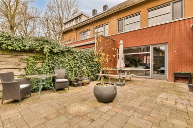 Sarmaşıklarla çevrili kırmızı tuğlalı bir evin önündeki tuğla duvarda sandalyeler, masalar ve bitkilerle dolu bir teras.