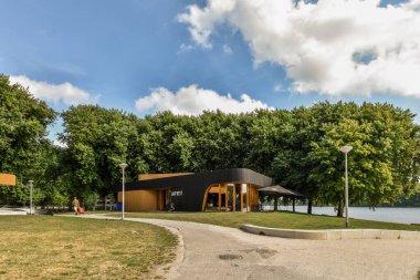 Amsterdam, Hollanda - 10 Nisan 2021: Bazı ağaçlarla dolu bir park ve gölün önündeki bankta oturan insanlar beyaz bulutlarla dolu açık mavi bir gökyüzünün altında