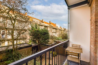 Arka planda ağaçlar ve binalar olan bir balkon. Bir apartmanın penceresinden sokağa bakıyor.