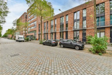 Amsterdam, Hollanda - 10 Nisan 2021: kaldırım boyunca büyüyen ağaçlar ve bitkilerle dolu bir cadde üzerinde bir apartmanın önüne park edilmiş iki araba