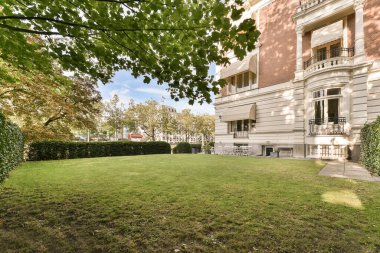 Büyük bir binanın önündeki çimenlikte kırmızı bir araba çimlerin üzerine park edilmiş ve arkasında ağaçlar var.