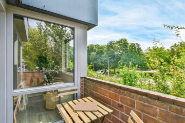 Güneşli bir günde, bir bankı, masası ve penceresi olan ve fotoğraftaki ağaçlara bakan bir dış alan çekilir.