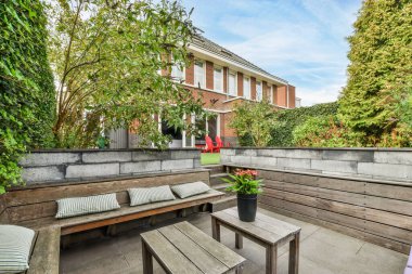 Ağaçlarla çevrili tuğla bir binanın önünde, dış duvarında ahşap banklar ve bitkiler olan bir teras.