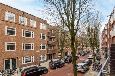 Amsterdam, Hollanda - 10 Nisan 2021: Birçok penceresi ve kapısı olan büyük bir tuğla binanın önündeki caddenin kenarına park edilmiş bazı arabalar