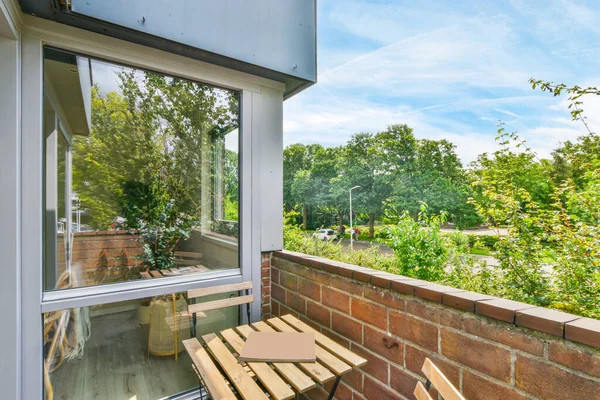 Güneşli bir günde, bir bankı, masası ve penceresi olan ve fotoğraftaki ağaçlara bakan bir dış alan çekilir.