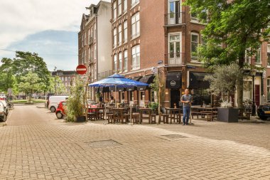 Bazı insanlar büyük tuğlalı bir binanın önündeki kaldırımda masalarda ve şemsiyelerde oturuyorlar.