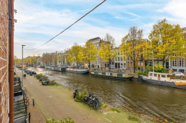 Amsterdam, Hollanda - 10 Nisan 2021: Nehrin diğer yakasında bazı tekneler ve arkasında ağaçlar olan binalar