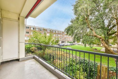 Balkonunda ağaçlar ve bitkiler olan bir dış alan. Apartman binalarının birinden sokağa bakıyor.