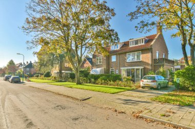 Amsterdam, Hollanda - 10 Nisan 2021: Her iki tarafında evler ve ağaçlar olan bir sokak, evin önünde park edilmiş yeşil bir araba var.