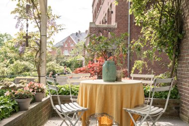 Masanın yanındaki vazolarda çiçekler olan bir verandada masa ve sandalyeler eski bir tuğla bina.