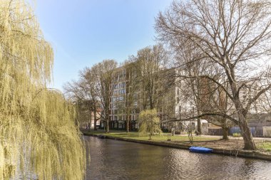 Amsterdam, Hollanda - 10 Nisan 2021: Arka planda ağaçlar ve nehrin diğer tarafında evler olan bir nehir, hepsi yapraksız dallarla çevrili.
