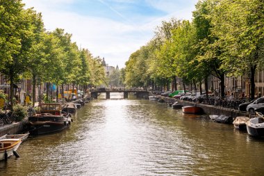 Amsterdam, Hollanda - 10 Nisan 2021: üzerinde birçok tekne bulunan bir kanal ve arkaplanda nehir kenarına dizili ağaçlar beyaz bulutlarla dolu bir gökyüzü.