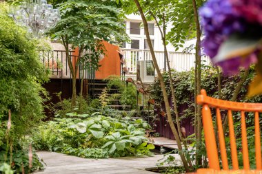 Amsterdam, Hollanda - 10 Nisan 2021: Ağaçlarla çevrili turuncu bir bank da dahil olmak üzere ön planda birçok bitki ve çiçeğin bulunduğu bir bahçe