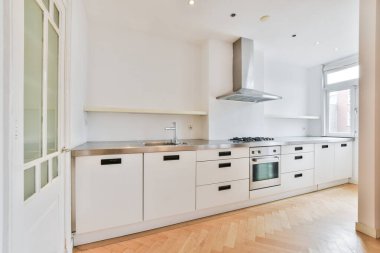 Fotoğrafın ortasında beyaz dolaplar ve ahşap döşemeler olan bir mutfak başka bir odaya açılan açık bir kapıdır.