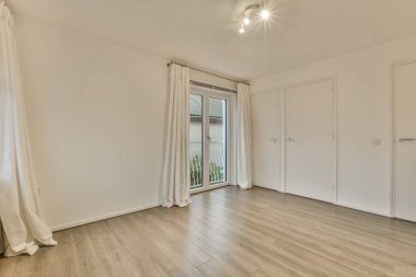 Beyaz duvarları ve ahşap döşemeleri olan boş bir oda, verandaya açılan sürgülü cam bir kapı var.