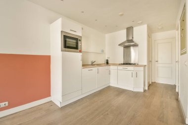 Odanın ortasında beyaz dolapları ve ahşap döşemeleri olan bir mutfak. Duvarda turuncu bir aksan var.