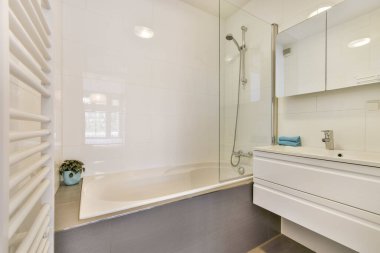 Beyaz duvarları ve gri zemini olan modern bir banyo. Lavabonun yanında serbest banyo küveti de var.