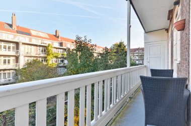 Ön tarafında sandalyeler ve ağaçlar olan bir balkon. Fotoğrafın sol tarafında pencereli bir alan var.