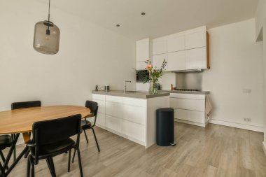 Beyaz duvarları, ahşap döşemeleri ve siyah sandalyelerle çevrili bir ada masası olan bir odada mutfak ve yemek odası.