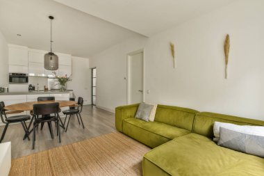 Salonda yeşil kanepesi ve yemek masası olan bir oturma odası beyaz duvarlar, ahşap döşemeler ve açık mutfak alanı var.