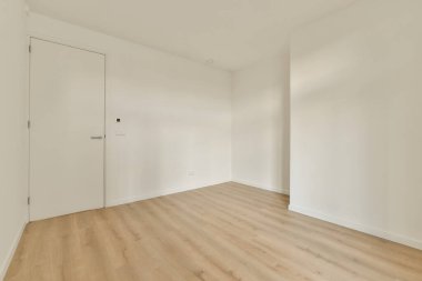 Beyaz duvarları ve sağ tarafında ahşap döşemeleri olan boş bir oda, sol köşede bir kapı var.