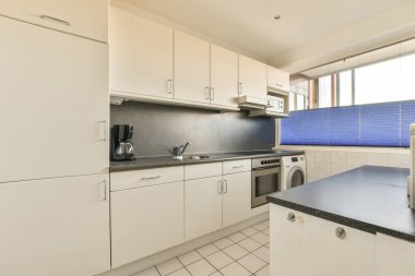 Bu fotoğrafta tezgahların üzerinde beyaz dolaplar ve siyah tezgahlar olan bir mutfak içeriden çekilmiş.