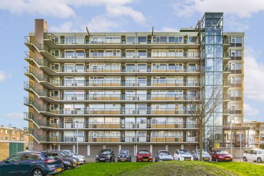 Otoparkta arabaların park ettiği bir apartman dairesi. Çimenli bir tepenin üzerinde. Etrafı yüksek binalarla çevrili.