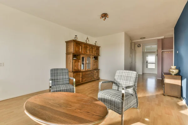 Ahşap döşemeleri ve beyaz duvarları olan bir oturma odası. İki mavi sandalyeyle çevrili ahşap bir sehpa da dahil.