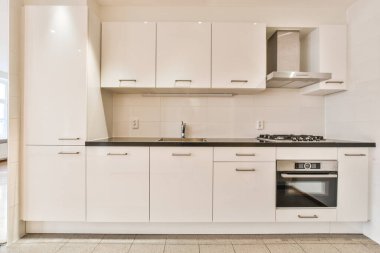 Bu fotoğrafta tezgahların üzerinde beyaz dolaplar ve siyah tezgahlar olan bir mutfak içeriden çekilmiş.