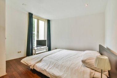 Ahşap döşemesi ve beyaz duvarları olan bir yatak odası, yatağın üstündeki duvarda bir televizyon var.