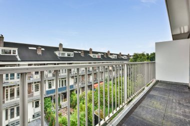 Beyaz parmaklıklı bir balkon ve yerde gri fayanslar var. Arka planda mavi bir gökyüzü var.