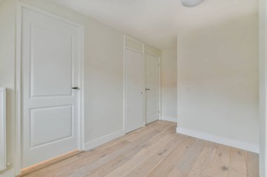 Ahşap zeminli boş bir oda ve duvarlarında beyaz kapılar olan başka bir odaya açılan bir kapının önünde.