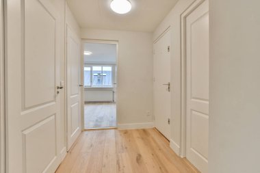 Odanın sağ tarafında beyaz duvarları ve ahşap döşemeleri olan boş bir oda, başka bir odaya açılan bir kapı var.